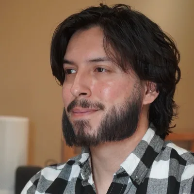 Brandon Delgado, smiling and looking aside, with wavy dark hair and a beard, in a black-and-white checked shirt.